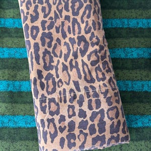 May include: A rectangular, folded fabric item with a brown and black leopard print pattern. The fabric is a light tan color with dark brown spots. The item is lying on a green and blue striped surface.