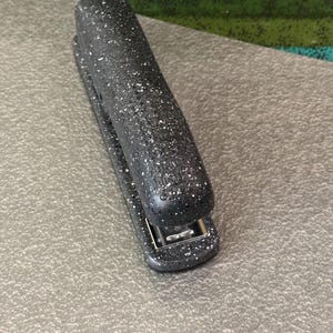 May include: A black stapler with a white speckled pattern across its exterior surface. The stapler features a rounded top design and a metallic silver mechanism visible at the front. The brand name Bostitch is embossed on the top of the stapler body.