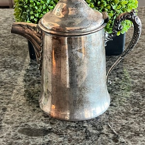 May include: A silver pitcher with a decorative handle and a lid with a rose finial. The pitcher has a tarnished finish and is sitting on a gray countertop.