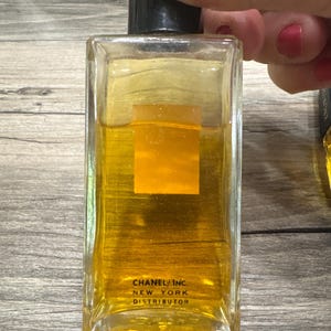 May include: A rectangular glass perfume bottle with a black cap, filled with a golden-colored liquid. The bottle has a square, orange-colored label and the text "CHANEL INC. NEW YORK DISTRIBUTOR" printed on the front.