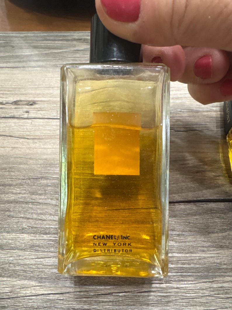 May include: A rectangular glass perfume bottle with a black cap, filled with a golden-colored liquid. The bottle has a square, orange-colored label and the text "CHANEL INC. NEW YORK DISTRIBUTOR" printed on the front.