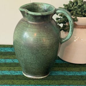 May include: A ceramic pitcher with a rounded body and a curved handle, featuring a mottled green and dark brown glaze with visible horizontal texture lines and a small pouring spout at the top rim of the vessel.