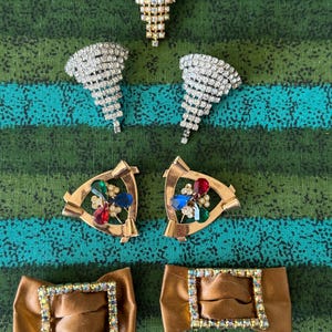 May include: A collection of vintage jewelry pieces, including three rhinestone brooches in silver and gold tones, two gold-toned earrings with colorful gemstones, and two brown satin bow clips with rhinestone borders.