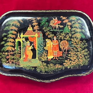 Puede incluir: Una bandeja lacada en negro con un borde dorado y una escena pintada de personas en un bosque. La escena incluye un castillo, árboles y flores. La bandeja está decorada con pan de oro y tiene una firma en la parte inferior.
