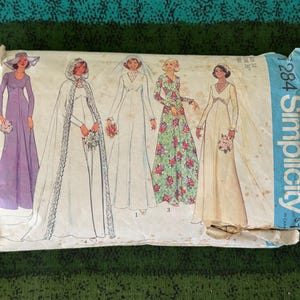 May include: A vintage Simplicity sewing pattern packet showing illustrations of five wedding dress designs. The designs include a long purple dress, a bridal gown with a cape, and a floral print dress. The packet is in a worn paper envelope.