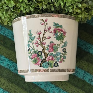 May include: A cream-colored ceramic planter featuring a central floral motif with pink, green, and blue blossoms on a brown branch. The top and bottom rims are decorated with a brown geometric border pattern, and the surface has a subtle crackle finish.