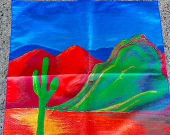 Puede incluir: Un tapiz colorido que presenta un paisaje desértico con un gran cactus verde en primer plano. El fondo muestra montañas rojas y verdes bajo un cielo azul.