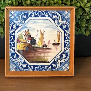 May include: A framed tile with a blue and white border depicting a Dutch seascape with sailboats and houses. The tile is painted in a traditional Delft blue style.