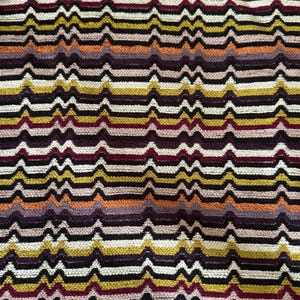 Può includere: Primo piano di un tessuto strutturato con un motivo a onde ripetute. Il design presenta strisce orizzontali in vari colori, tra cui bordeaux, oro, bianco, nero, arancione e viola. Il tessuto ha un aspetto morbido e tessuto.