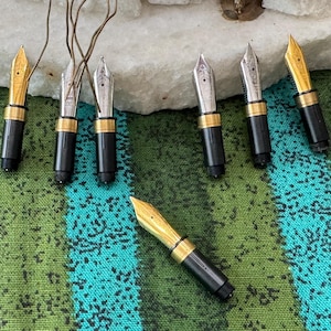 May include: A collection of seven fountain pen nib units, each featuring a black plastic grip section and a metallic nib. Three nibs are gold-toned, and four are silver-toned. The nibs are arranged on a green and teal striped fabric surface.