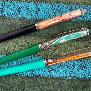 Op de afbeelding: Drie intrekbare balpennen met doorzichtige houders met zwevende afbeeldingen. De bovenste pen heeft een zwarte grip en een brugillustratie. De middelste pen heeft een groene grip en een groene afbeelding. De onderste pen heeft een groenblauwe grip en een oranje afbeelding.