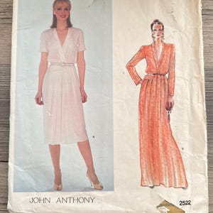 May include: Vintage Vogue American Designer sewing pattern featuring two dress designs. One dress is a short-sleeved wrap dress with a belt, and the other is a long-sleeved, floor-length dress. The pattern includes the text "JOHN ANTHONY" and "2522".