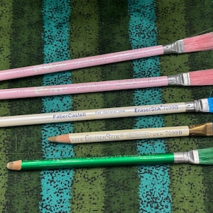 May include: Four pencils with brush-style erasers. Two are pink with pink erasers, one is white with a blue eraser, and one is green with a green eraser. The pencils have text that reads "Faber Castell" and "EraserStik 7099B".