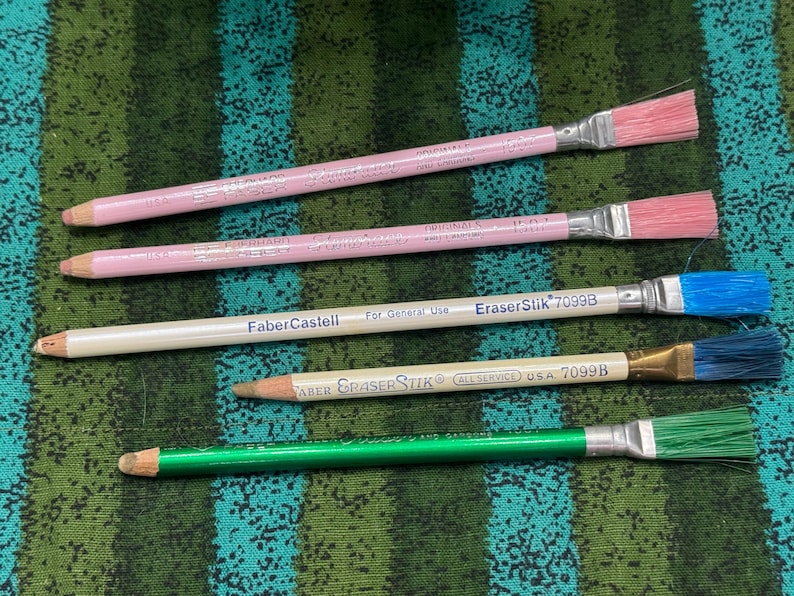 May include: Four pencils with brush-style erasers. Two are pink with pink erasers, one is white with a blue eraser, and one is green with a green eraser. The pencils have text that reads "Faber Castell" and "EraserStik 7099B".
