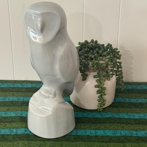 May include: A smooth, light gray ceramic owl figurine standing on a cylindrical base. The owl has a heart-shaped facial disc and a minimalist, stylized form with a glossy finish, positioned next to a small white pot containing a green succulent plant.