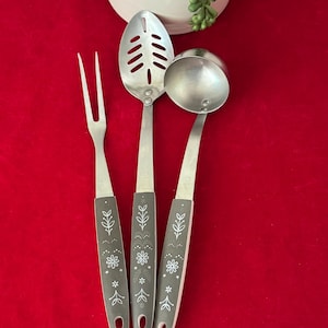 May include: A set of three stainless steel kitchen utensils: a fork, a slotted spoon, and a ladle. Each utensil has a gray handle with a white floral design. The utensils are arranged on a red surface.