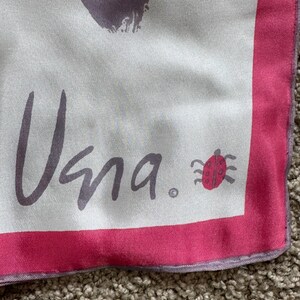 May include: A square silk scarf with a white background and a pink border. The scarf features the word "Vana" in gray script, along with a small red ladybug illustration. The design includes abstract purple brushstrokes.