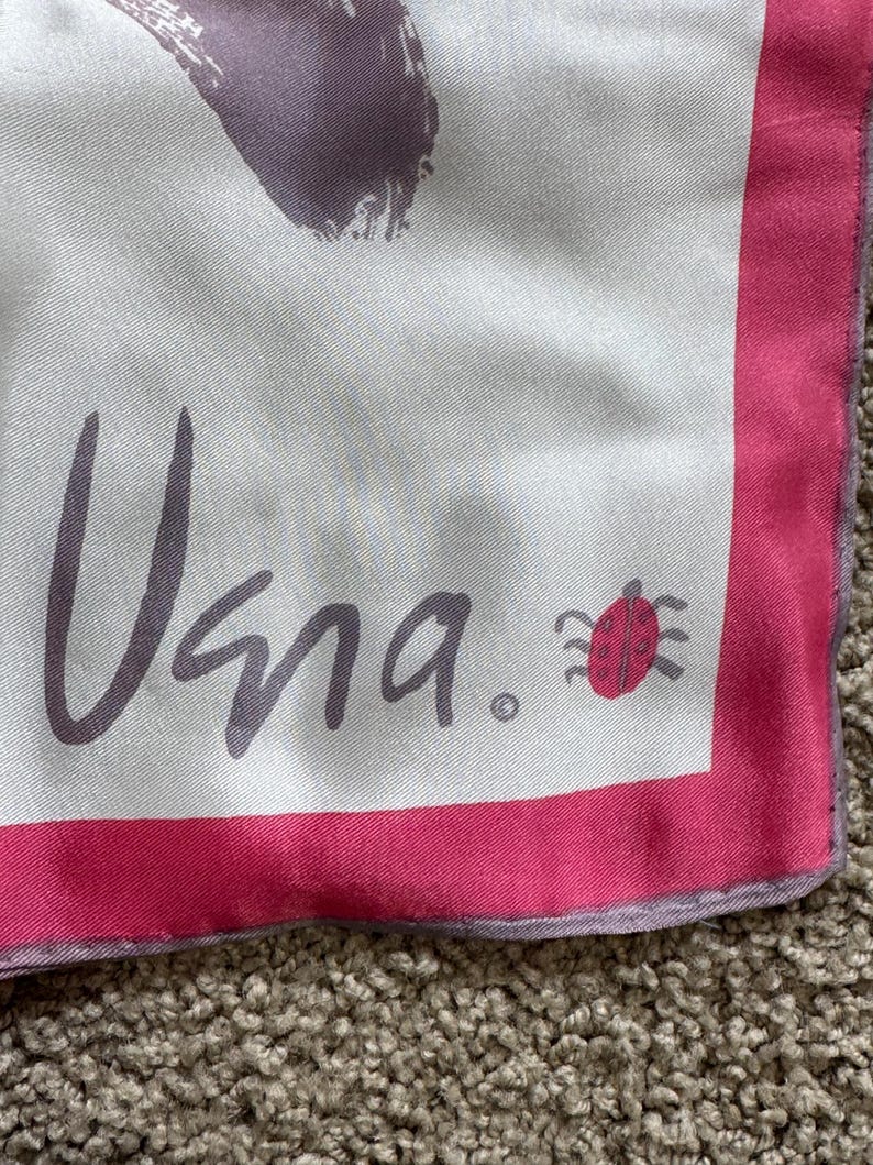 May include: A square silk scarf with a white background and a pink border. The scarf features the word "Vana" in gray script, along with a small red ladybug illustration. The design includes abstract purple brushstrokes.