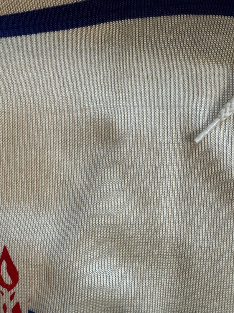 May include: Close-up of a beige knitted fabric with a blue and white stripe at the top. The fabric has a subtle texture and a slight sheen.