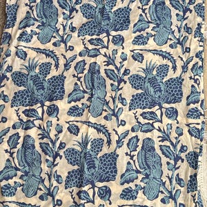 May include: A fabric with a repeating pattern of blue birds and floral designs on a cream background. The birds are perched on branches with leaves and flowers. The fabric has a vintage or bohemian aesthetic.