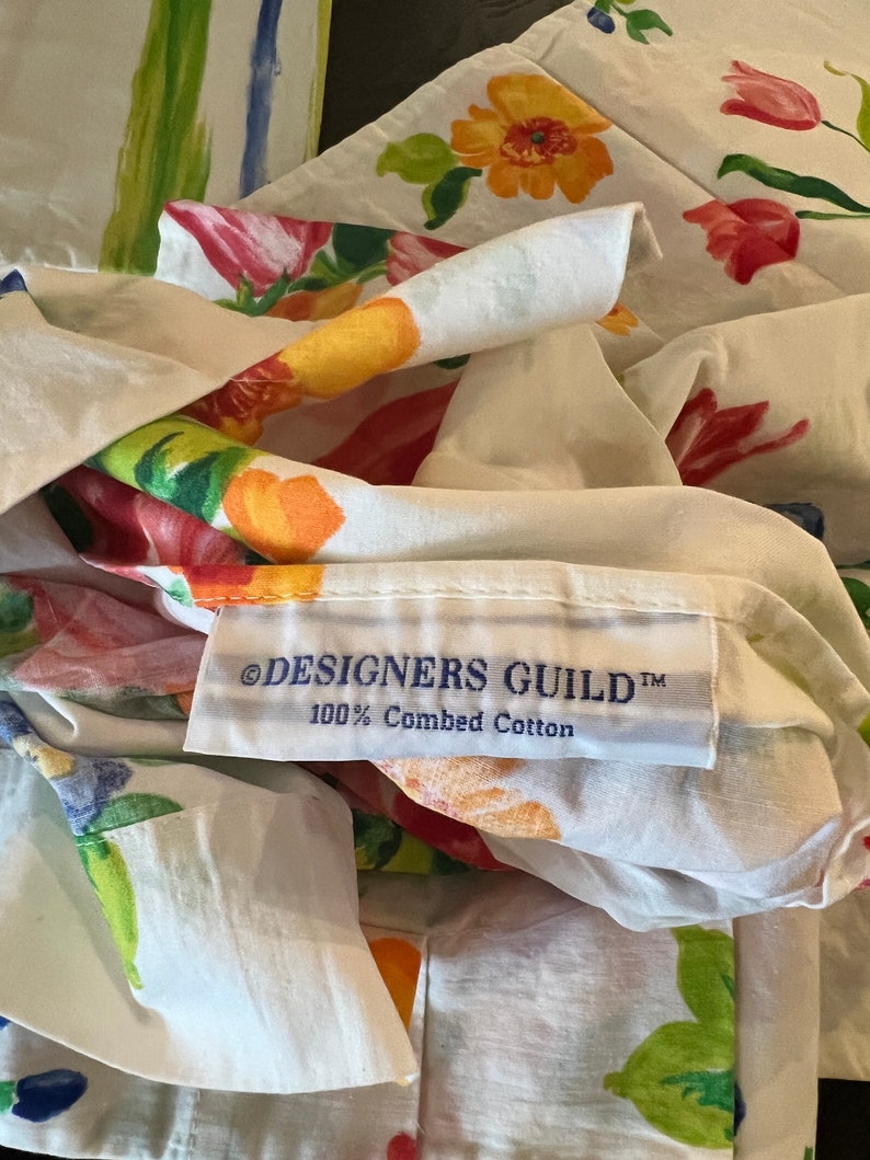 May include: White fabric with a floral print of tulips, leaves, and flowers in shades of red, orange, yellow, green, and blue. The fabric has a label that reads "&copy;DESIGNERS GUILD&trade; 100% Combed Cotton".