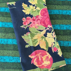 May include: Folded navy blue floral print fabric featuring large pink and red roses, green leaves, and clusters of pink grapes. The fabric has a green border and is set against a striped background.