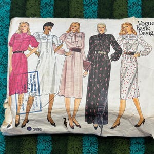 Op de afbeelding: Vintage Vogue Basic Design naaipatroon envelop met illustraties van vijf verschillende jurken. De jurken variëren in kleur en stijl, met details zoals riemen, strikken en bloempatronen. De envelop is licht versleten, met het nummer 2896 zichtbaar.