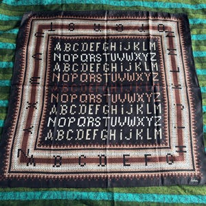 Peut inclure: Un foulard carré noir avec un motif d'alphabet répété en blanc. Le foulard a une bordure marron et beige avec un motif géométrique répétitif. Le foulard est fait d'une matière soyeuse et est plié sur une surface rayée verte et bleue.