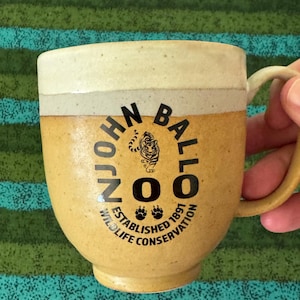 May include: A ceramic mug with a speckled tan and cream glaze. The front features black text reading JOHN BALLO ZOO, an illustration of a tiger, and the words ESTABLISHED 1891 WILDLIFE CONSERVATION with two paw print icons below the tiger.
