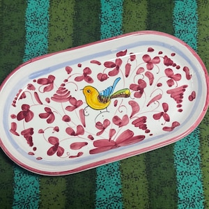 May include: An oval ceramic tray with a white base, featuring a hand-painted yellow bird with blue and green wing accents at the center. The tray is decorated with scattered magenta floral motifs and has a light blue inner rim and a pink outer border.
