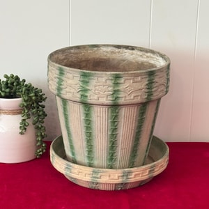 May include: A large, vintage-style ceramic flower pot with a matching saucer. The pot is beige with vertical green stripes and decorative green trim. The pot sits on a red surface.