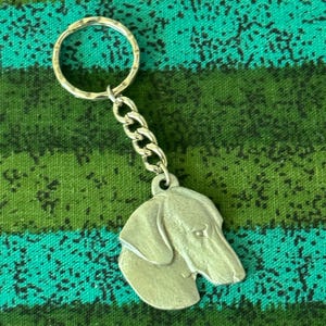 May include: A silver-toned metal keyring featuring a sculpted dog head charm attached to a circular split ring by a short chain. The charm depicts the profile of a dog with floppy ears and a long snout, rendered in a matte, brushed metal finish.