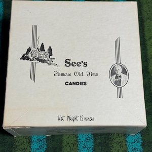 May include: A white box of See's Famous Old Time CANDIES. The box features a black and white illustration of a house and trees, along with a portrait of a man. The net weight is 12 ounces.