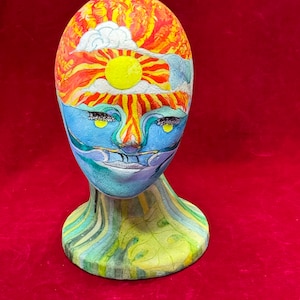 May include: A ceramic head sculpture with a painted design of a sun, clouds, and waves. The head is on a green and yellow striped base.