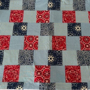 May include: A patchwork quilt featuring a repeating pattern of red paisley squares, navy blue floral squares, light blue gingham squares, and solid light blue squares. The quilt is made of fabric and has a textured appearance.