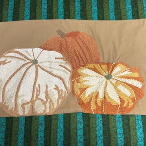 May include: A rectangular tan pillow cover featuring three embroidered pumpkins. One large pumpkin is white with tan accents, one is solid orange, and the third is a smaller orange and white pumpkin. The embroidery has a textured, raised appearance.