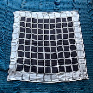May include: A square scarf with a black and white checkered pattern. The scarf has a border of white squares and a center of black squares. The scarf is made of a silky material and is laying on a blue surface.
