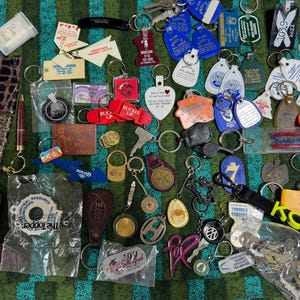 May include: A large collection of assorted keychains and key rings in various shapes, colors, and materials, including plastic, metal, and leather. The items feature diverse logos, text, and designs, with some items still in their original plastic packaging.