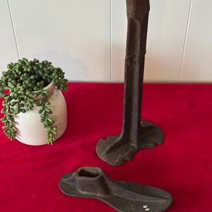 May include: Two antique cast iron shoe lasts. The larger last is a shoe stand with a tall, narrow base and three legs. The smaller last is a shoe form with a rounded toe and a hollowed-out heel.