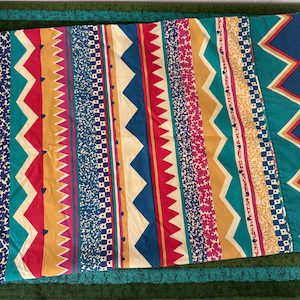 May include: A rectangular fabric textile featuring a repeating vertical pattern of multicolored stripes. The design includes bands of teal, mustard yellow, magenta, and navy blue, accented with zigzag lines, small floral motifs, and checkered squares.