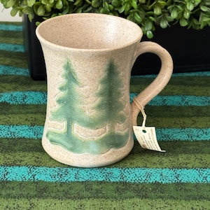 May include: A beige ceramic mug with a green tree design. The mug has a curved handle and a slightly flared rim. The trees are painted in a muted green colour. A small tag is attached to the handle.