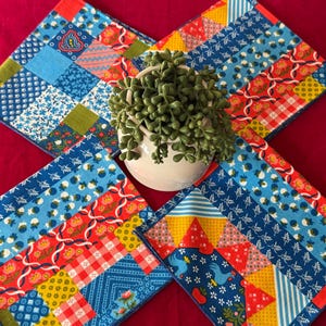 Puede incluir: Cuatro posavasos cuadrados de tela colorida con patrones de patchwork en azul, rojo, amarillo y verde. Una pequeña maceta de cerámica blanca con una planta verde se encuentra en el centro. Los posavasos tienen varios diseños florales y geométricos.