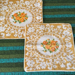 May include: Two square terry cloth towels featuring a white floral pattern on a mustard yellow background. Each towel has a central white circular medallion containing a cluster of orange roses with green leaves and stems.