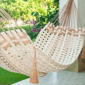 May include: A woven hammock in cream and tan, hanging outdoors. The hammock features a woven pattern with rectangular openings and a decorative tassel. The background includes green foliage and a glimpse of a white structure.