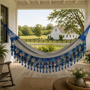 May include: A gray hammock with blue and white floral accents and tassels hangs on a porch. A wooden rocking chair with a striped pillow sits nearby. The background features a white farmhouse and green landscape.