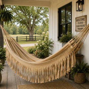 May include: A beige hammock with a fringed edge hangs on a porch. The hammock is suspended between wooden posts. A sign that reads "Welcome to our Farm House" is visible. Potted plants and a rug complete the outdoor scene.