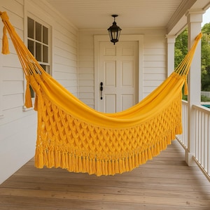 Puede incluir: Una hamaca de color amarillo brillante con un fleco decorativo tejido al estilo macramé a lo largo del borde inferior. La hamaca está suspendida por gruesas cuerdas trenzadas en ambos extremos, con un cuerpo de tela sólida.