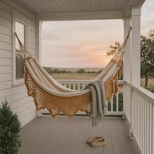 May include: A cream-colored fabric hammock with mustard yellow macramé fringe and tassels suspended on a porch. A gray knit throw blanket is draped over the edge of the hammock, and a pair of beige slip-on shoes sits on the floor beneath it.