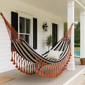 May include: A black and white striped hammock with orange rope and tassels hangs on a porch. A white pillow with a striped pattern rests inside the hammock. The hammock is suspended between white columns.