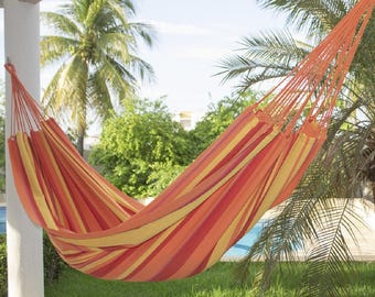 May include: A fabric hammock featuring a vibrant striped pattern in shades of orange, yellow, and red. The hammock is suspended by thick, matching orange cords attached to a white pillar, showcasing a woven design with a deep, curved center.